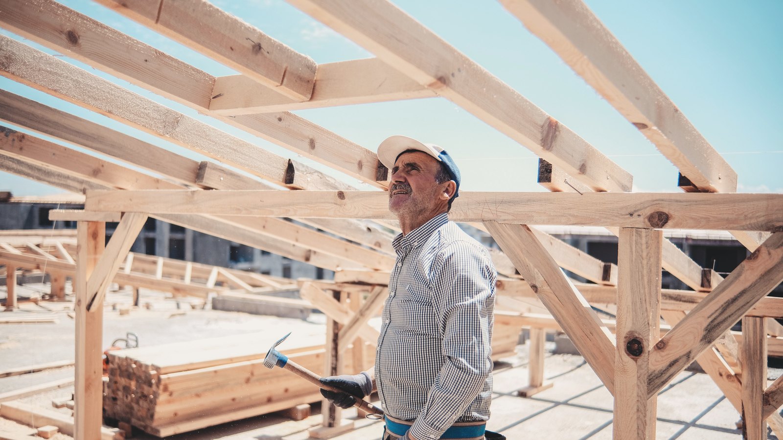 A tradesman under a bare roof frame looks up at the work, hammer resting at his side